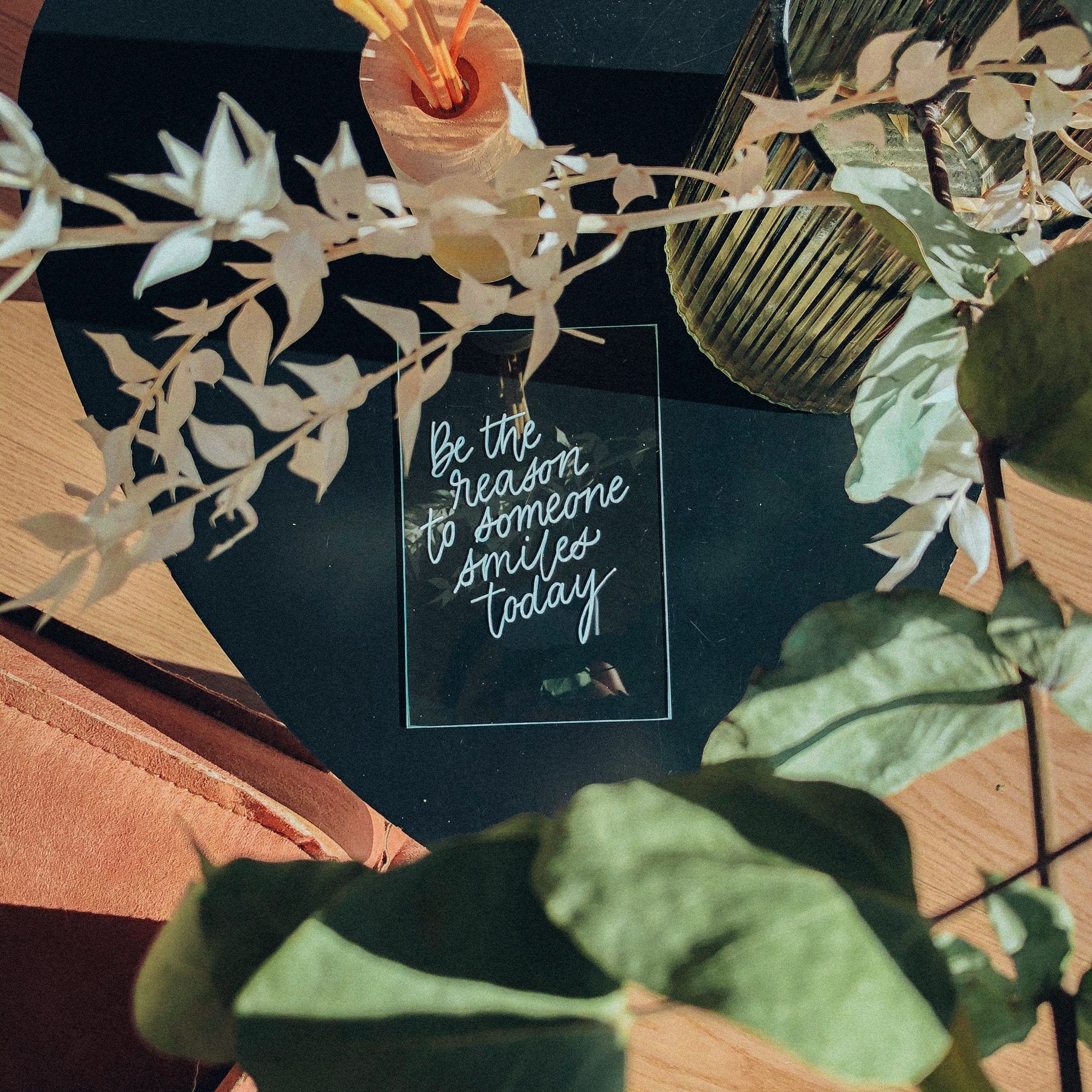 Panneau en verre calligraphié « Be the reason someone smiles today », entre des feuillages
