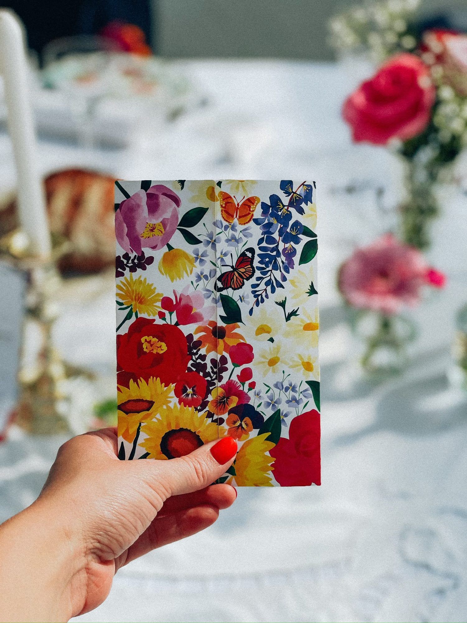 Faire-part de mariage aux fleurs peintes et papillons, tenu à la main devant une table fleurie
